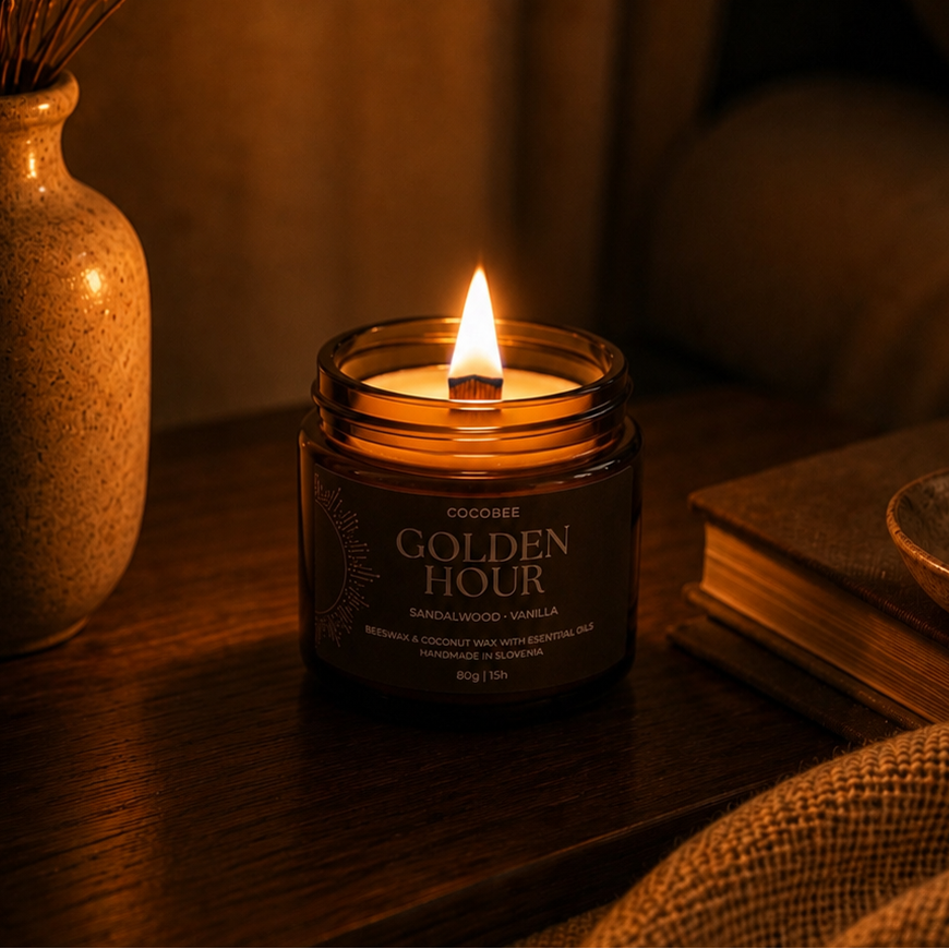 A lit candle in a dark amber glass jar sits on a wooden table in a cozy room setting, with a vase of dried stems on the left and a book on the right. The label reads 'GOLDEN HOUR'.