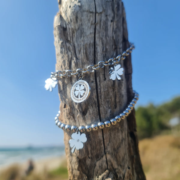 Pulsera de acero con cadena rolo que presenta 2 tréboles de cuatro hojas colgantes y 1 trébol central con piedras blancas