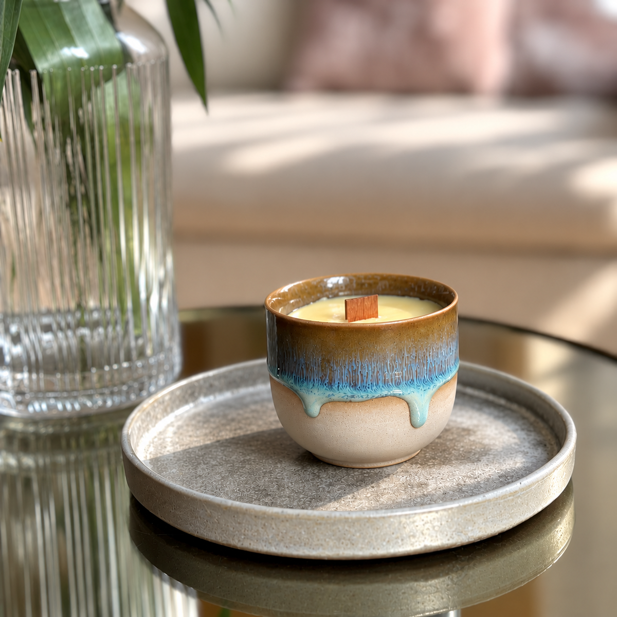 A small ceramic candle jar with a brown and blue glazed drip design sits on a round gray tray on a reflective table, with a glass vase and softly blurred sofa in the background.