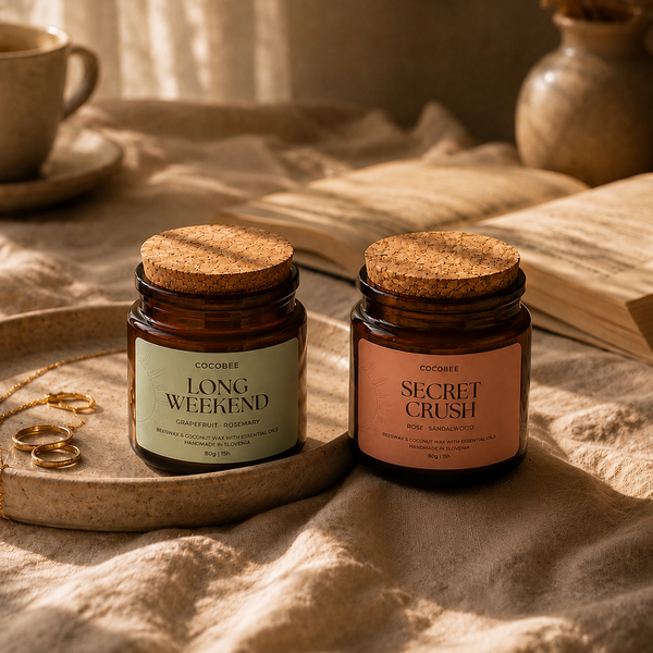Two amber glass jar candles with cork lids on a draped beige fabric surface. One label reads 'LONG WEEKEND' and the other reads 'SECRET CRUSH'.