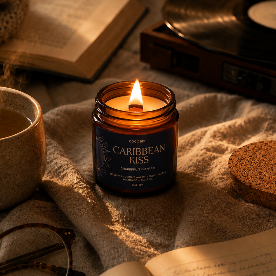 A lit amber glass jar candle labeled 'COCONESS CARIBBEAN KISS' sits on a beige knit blanket with a mug, glasses, an open notebook, and warm cozy props around it.