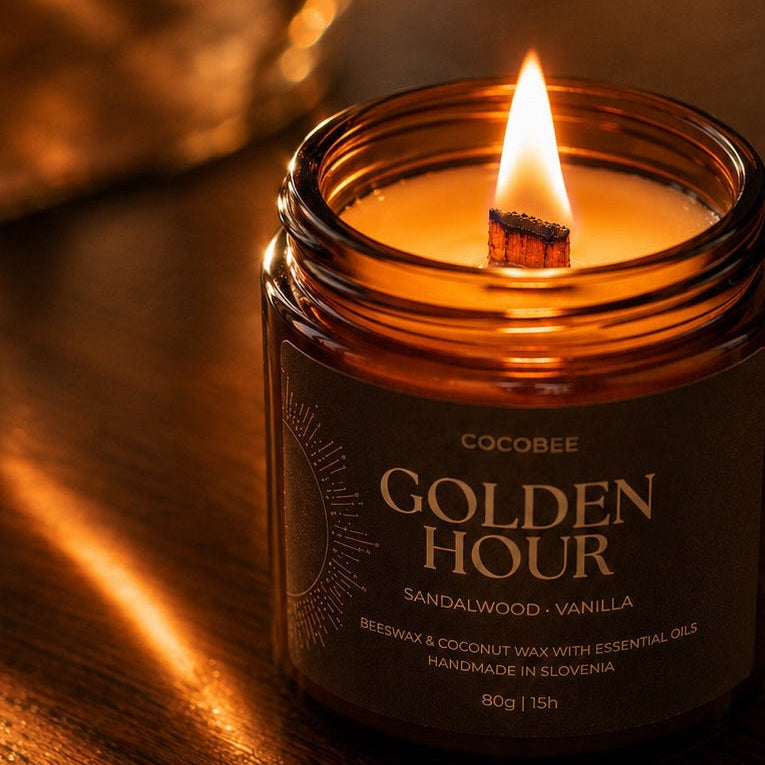 A lit Cocosbee Golden Hour candle in an amber glass jar sits on a dark wooden table, casting a warm glow. The label reads 'GOLDEN HOUR - SANDALWOOD - VANILLA'.