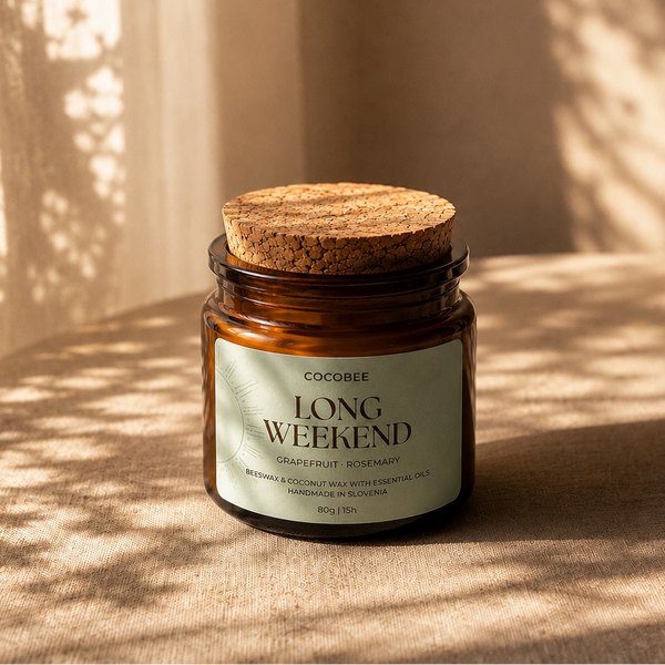An amber glass jar candle with a cork lid and a cream label reading 'COCOREE LONG WEEKEND' sits on a sunlit beige surface with warm shadows cast across the scene.