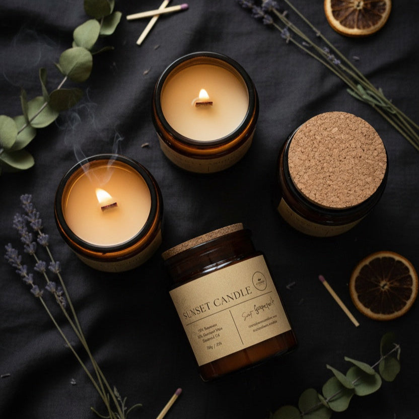 Two lit candles in brown jars with cork lids on a dark surface with dried lavender and oranges. Sunset Candle in amber glass jar with crackling wooden wick. Beeswax & Coconut wax.