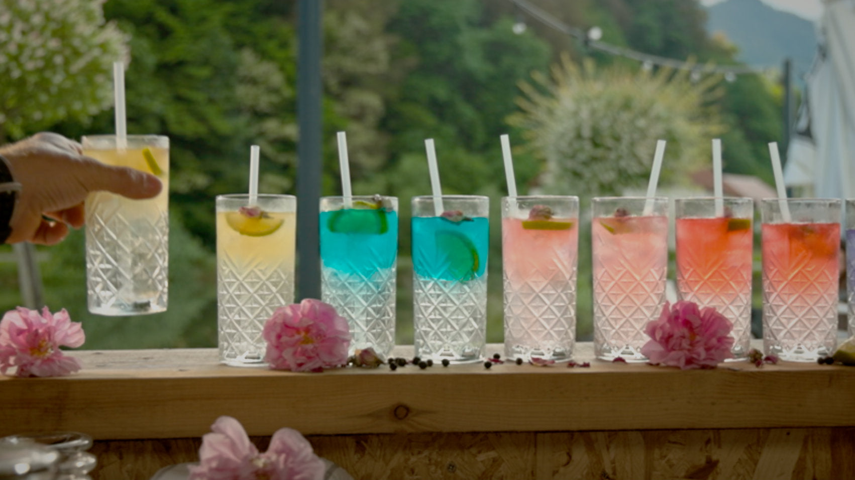 Hand reaching to grab a tall, clear cocktail glass among a row of colorful drinks on a sunlit wooden bar, outdoors.