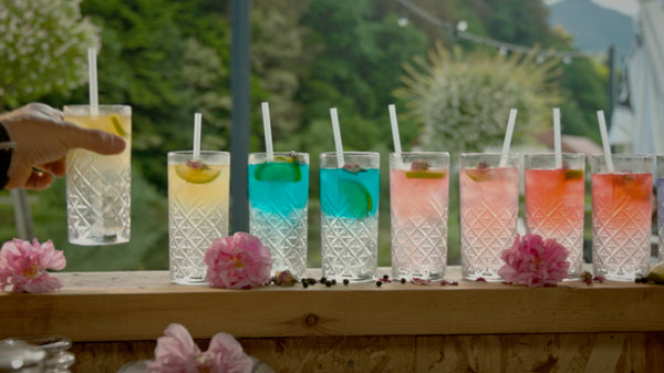 Hand reaching to grab a tall, clear cocktail glass among a row of colorful drinks on a sunlit wooden bar, outdoors.