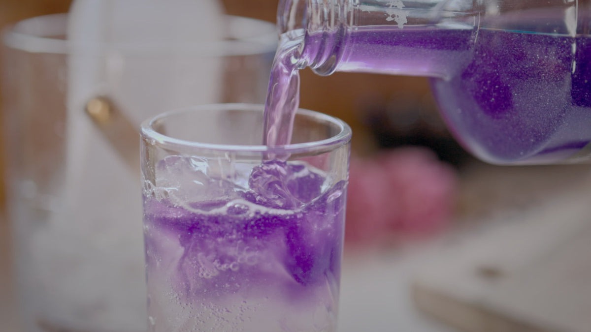 Purple beverage being poured into a tall glass over ice cubes.