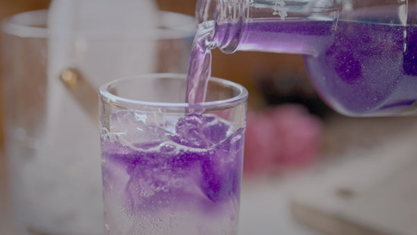 Purple beverage being poured into a tall glass over ice cubes.