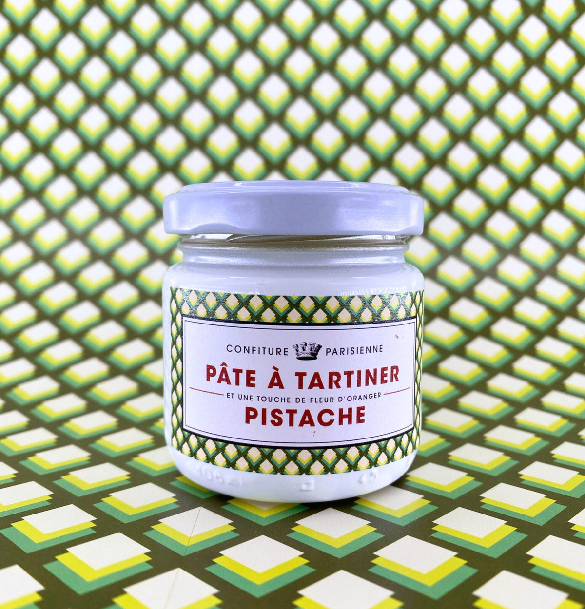 Pot de Pâte à Tartiner Pistache et fleur d'oranger posé sur un fond graphique à motif d'écailles vert et jaune