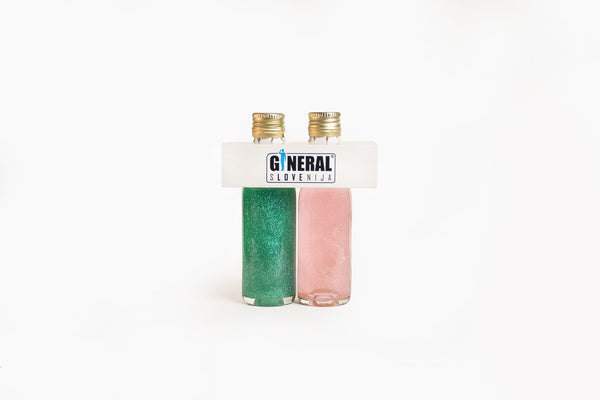 Two glitter nail polish bottles, green and pink, standing side by side with a white carrier labeled 'GENERAL SLOVENIJA' on top.