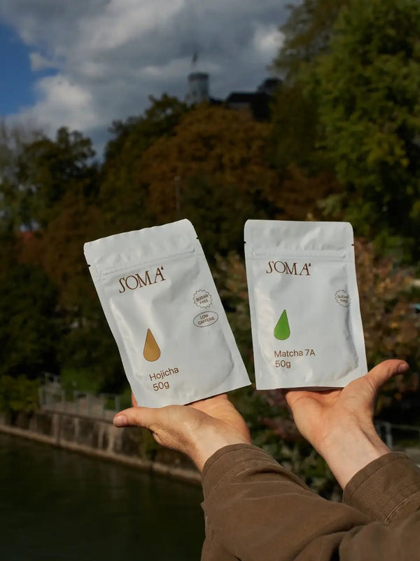 Hands holding Soma Hojicha and Matcha 7A pouches by a river