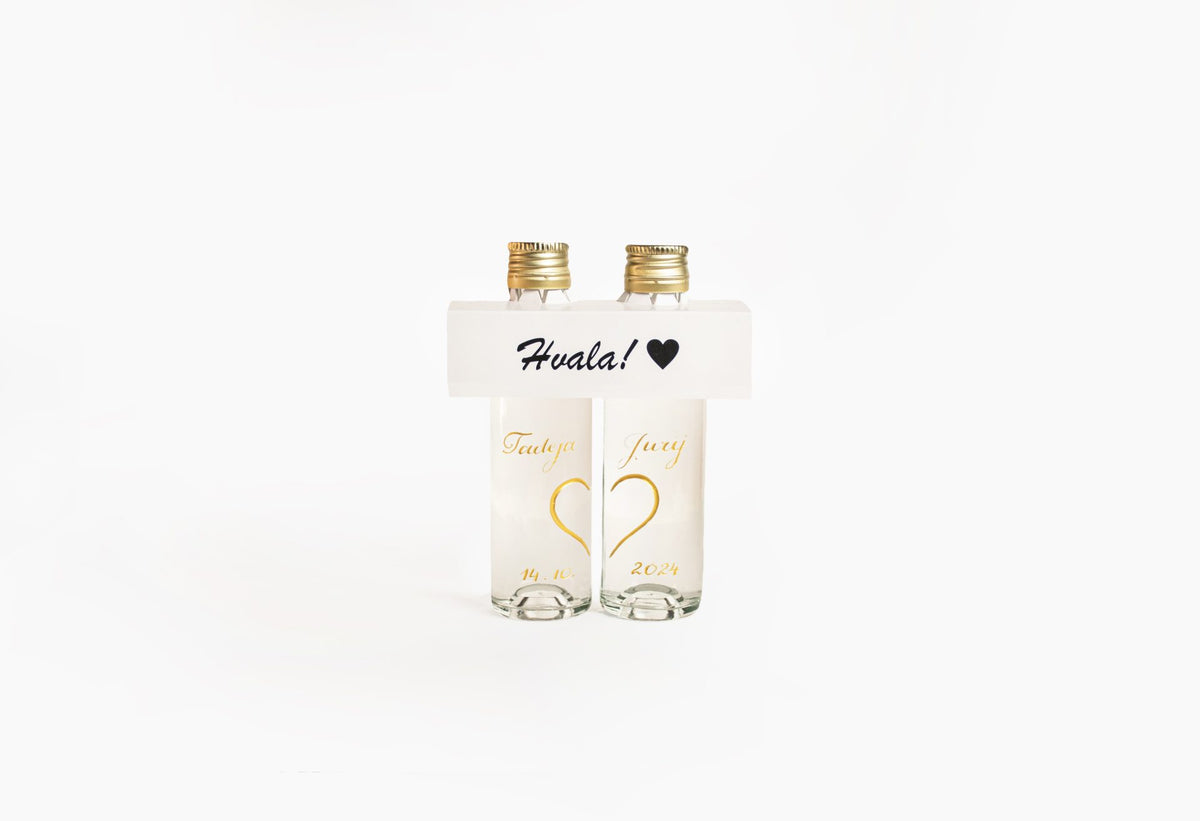 Two clear bottles with gold caps and white labels displaying gold heart designs; a white band across the top reads 'Hvala!' with a black heart symbol.