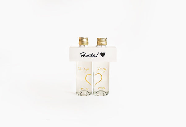 Two clear bottles with gold caps and white labels displaying gold heart designs; a white band across the top reads 'Hvala!' with a black heart symbol.